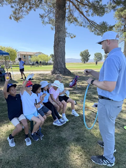Kids on a bench under a pine tree raise their hands as Coach Jon leads a junior clinic