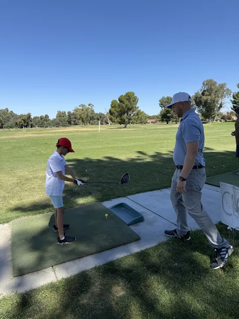 A young golfer in a red cap lines up a teed ball as Coach Jon watches