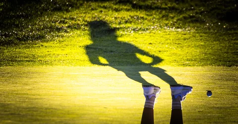 A golfer’s long shadow across a golden fairway, beside the ball